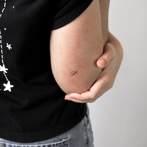 May include: A black t-shirt featuring a white graphic of stars connected by dotted lines and a small, delicate black script letter P tattoo located on the skin near the elbow. The garment is paired with grey denim fabric visible at the bottom.