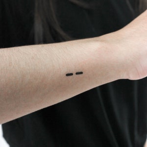 May include: A close-up of a person's forearm with a small black tattoo of an em dash.