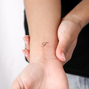 May include: A small, black, temporary tattoo of the cursive letter F applied to the skin of a wrist. The letter is rendered in a delicate, flowing script font with a thin line weight, positioned centrally on the inner forearm area.