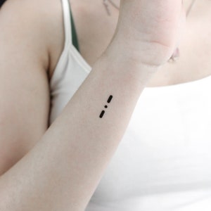 May include: A close-up of a person's wrist with a small black semicolon tattoo.