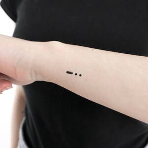 May include: A close-up of a person's arm with a small black tattoo of a dash and two dots.