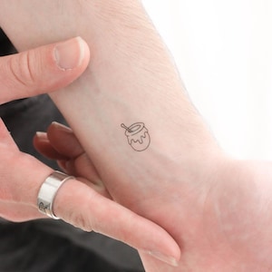 Puede incluir: Un tatuaje simple de línea de un tarro de miel con un cucharón en la muñeca de una persona.