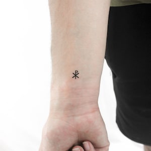 Könnte beinhalten: Ein kleines schwarzes Tattoo des Chi-Rho-Symbols auf dem Handgelenk einer Person.