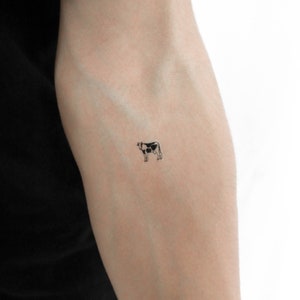 Può includere: Un piccolo tatuaggio di una mucca bianco e nero sul braccio di una persona.