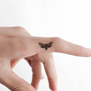 Könnte beinhalten: Ein schwarz-weißes temporäres Tattoo eines Nachtfalters auf einem Finger.