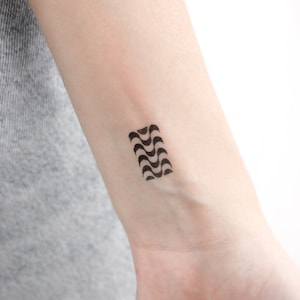 May include: A black and white geometric tattoo design on a person's wrist. The design is a series of curved lines that create a wave-like pattern.