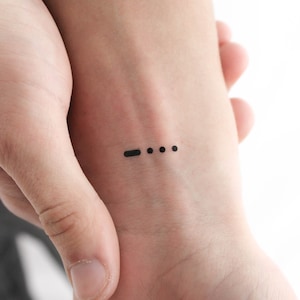Könnte beinhalten: Ein schwarzes Tattoo auf dem Handgelenk einer Person. Das Tattoo ist ein Strich, gefolgt von drei Punkten.