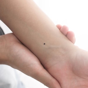 May include: A close-up of a person's wrist with a small black dot tattoo.
