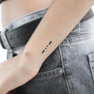 May include: A simple black tattoo of three dots on a person's wrist.