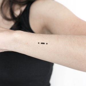Op de afbeelding: Een close-up van iemands arm met een kleine zwarte tijdelijke tatoeage van drie stippen, die de morsecode voor '…' (wat 'stop' betekent) voorstelt.