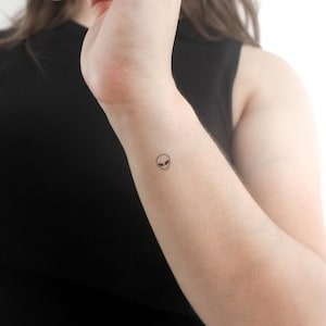 Può includere: Un piccolo tatuaggio semplice di un viso alieno sul polso di una persona.