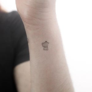 May include: A simple line drawing tattoo of a coffee pot on a person's wrist.