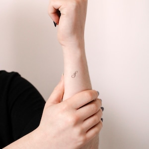 May include: A small, black, fine-line temporary tattoo of the cursive letter S positioned on the inner wrist. The design features a delicate, flowing script style with thin, precise lines, creating a minimalist and clean appearance on the skin.