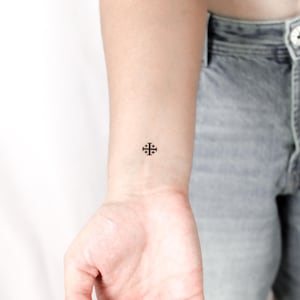 May include: A tiny black Jerusalem cross tattoo on a wrist. The design consists of a large cross with four smaller crosses inside, a Christian symbol. The tattoo is minimalist, with clean lines and a simple aesthetic, a popular choice.