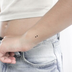 May include: A close-up of a person's wrist with a small tattoo of two black dots.