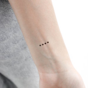May include: A close-up of a person's wrist with a small tattoo of four black dots in a row.