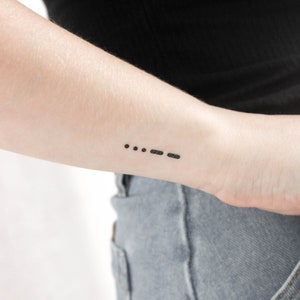 May include: A close-up of a person's wrist with a small black tattoo of three dots and three dashes, commonly known as Morse code for the letter 'S' and the number 'O'.