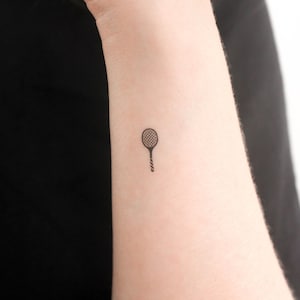 May include: A small black and white tennis racket tattoo on a person's wrist.