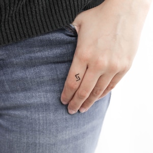 Può includere: Un piccolo tatuaggio temporaneo nero a forma di fulmine sul dito di una persona.