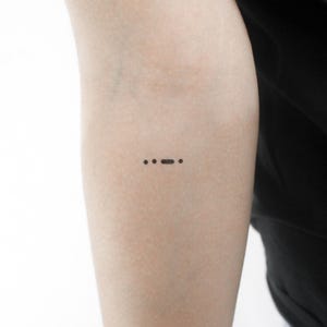 May include: A close-up of a person's arm with a small tattoo of three dots and a dash, representing the Morse code for the letter 'S'.
