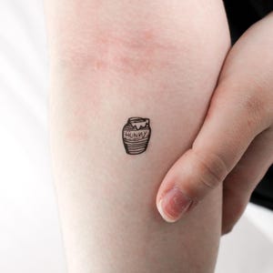 Puede incluir: Un pequeño tatuaje en blanco y negro de un tarro de miel con la palabra "HUNNY" en la parte delantera. El tarro tiene tapa y está lleno de miel. El tatuaje está en el brazo de una persona.