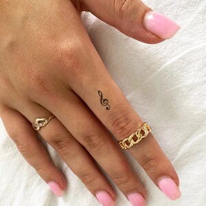 May include: A close-up of a hand with a small treble clef tattoo on the finger. The hand is wearing a gold chain ring.