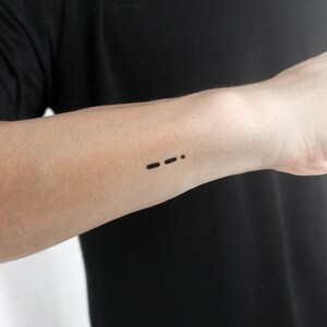 May include: A close-up of a person's arm with a small black tattoo of three dots and a dash, resembling Morse code.