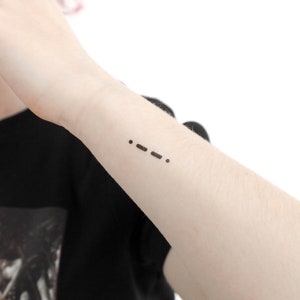 May include: A close-up of a person's wrist with a small tattoo of three dots in Morse code, representing the phrase "..."