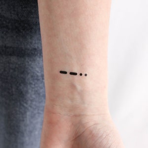 May include: A black tattoo on a person's wrist. The tattoo is a dash and three dots, representing Morse code for the letter 'S'.