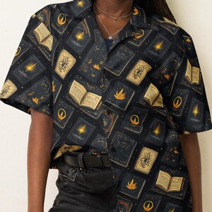 May include: A black button-down shirt with a short sleeve and a pattern of gold books and stars. The shirt is worn by a person with black hair and a black hat.