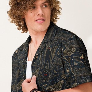 May include: A short-sleeved button-down shirt in dark navy blue featuring a repeating pattern of winged moth-like creatures, bridges, street lamps, and a central dark silhouette figure with glowing red eyes, worn over a plain white undershirt.
