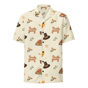 May include: A beige short-sleeved button-up shirt with a Thanksgiving theme. The shirt features a repeating pattern of turkeys, pilgrim hats, and the text "thanks giving".