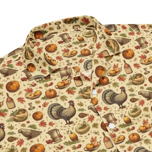 May include: A beige button-up shirt with a repeating pattern of fall harvest imagery. The pattern includes turkeys, pumpkins, gourds, leaves, and other fall harvest symbols.