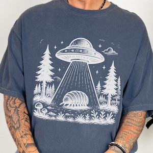 May include: A dark blue t-shirt with a white graphic of a UFO hovering over a pill bug in a forest setting. The graphic includes stars and a cloud.