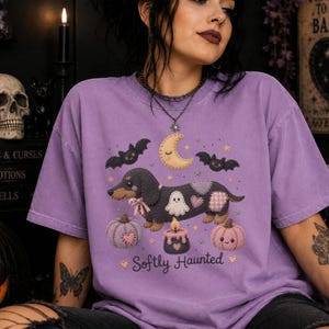 Può includere: Una maglietta lilla con un design a tema Halloween. La grafica include un bassotto, pipistrelli, una luna crescente, zucche, un fantasma e una candela. È presente anche il testo "Softly Haunted". Il design è in stile cartone animato.