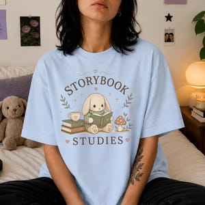 Puede incluir: Una camiseta azul claro con un gráfico de un conejo beige con un suéter verde leyendo un libro. El diseño incluye una pila de libros, una taza de té, un hongo y el texto "STORYBOOK STUDIES" en marrón, rodeado de pequeños corazones.