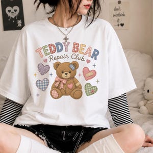 May include: White t-shirt with a graphic of a brown teddy bear with a pink bow, band-aid, and hearts. The text "TEDDY BEAR Repair Club" is above the bear. The shirt has a layered look with a black and white striped long sleeve underneath.