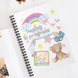 May include: A white spiral bound notebook with a rainbow, clouds, stars, a teddy bear, a baby bottle, a pacifier, and a book with a crescent moon on the cover. The text on the notebook says "healing in progress (please be kind)".