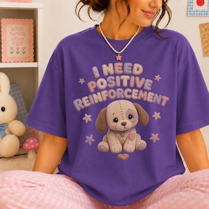 May include: A purple short-sleeve t-shirt featuring a graphic of a tan puppy with stitched details. The text "I NEED POSITIVE REINFORCEMENT" is printed above the puppy in a textured, multi-colored font, surrounded by small star and heart shapes.