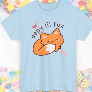May include: A light blue t-shirt featuring a cartoon fox illustration. The orange fox is curled up with a white-tipped tail. The text "eepy lil fox" and small pink hearts are above the fox.