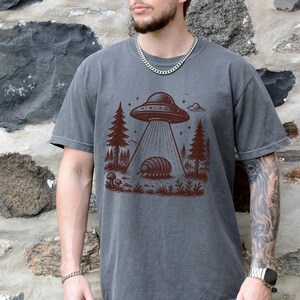 May include: A dark gray t-shirt with a graphic print of a flying saucer hovering over a pill bug in a forest setting. The graphic is in brown ink.