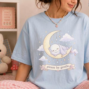 Pode incluir: Camiseta azul clara com uma ilustração de um personagem dormindo em uma lua crescente, cercado por estrelas e nuvens. O design inclui o texto "please be gentle" em uma faixa. A camiseta tem um corte folgado.