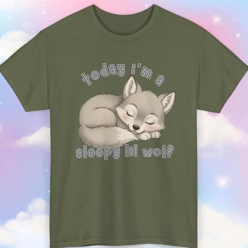 May include: Olive green t-shirt featuring a sleeping cartoon wolf illustration. The wolf is gray with pink accents, and the text "today i'm a sleepy lil wolf" is above and below the wolf. The shirt is made of a soft material.