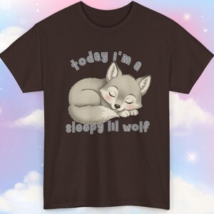 May include: A brown t-shirt featuring a sleeping cartoon wolf in shades of gray, with closed eyes and a pink nose. The text "today i'm a sleepy lil wolf" is arched above and below the wolf illustration.
