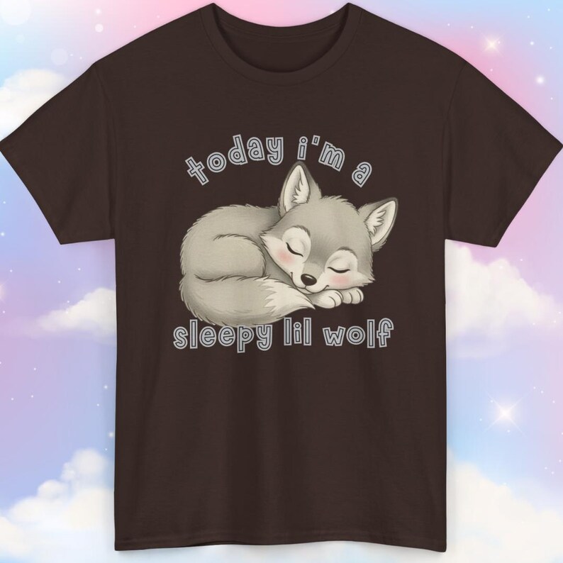 May include: A brown t-shirt featuring a sleeping cartoon wolf in shades of gray, with closed eyes and a pink nose. The text "today i'm a sleepy lil wolf" is arched above and below the wolf illustration.