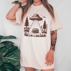 May include: A white t-shirt with a graphic of a UFO hovering over a pillbug in a forest setting. The graphic is in brown ink and features a starry sky.