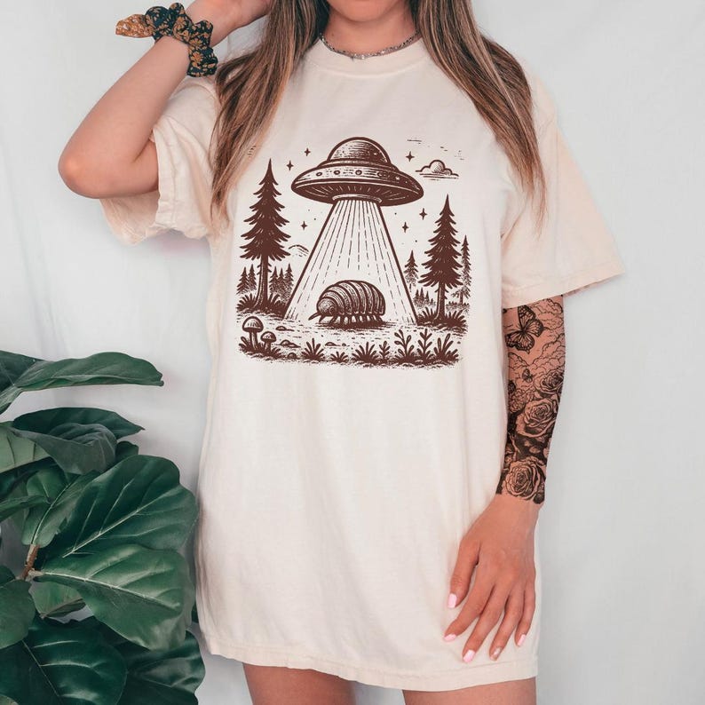 May include: A white t-shirt with a graphic of a UFO hovering over a pillbug in a forest setting. The graphic is in brown ink and features a starry sky.