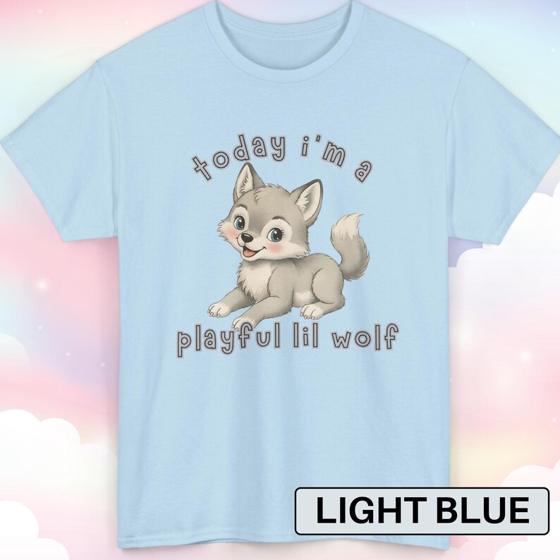 以下が含まれることがあります： 漫画のオオカミのデザインが施された水色のTシャツ。オオカミはグレーと白で、ピンク色のほっぺをしており、テキストは「today i'm a playful lil wolf」と書かれています。Tシャツには「LIGHT BLUE」と書かれています。