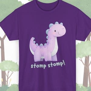 Op de afbeelding: Paars T-shirt met een cartoon dinosaurus in roze en blauw. De dinosaurus heeft een vriendelijke uitdrukking en de woorden "stomp stomp!" staan eronder. Het shirt is gemaakt van zacht materiaal.