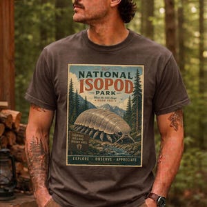 Op de afbeelding: Een antracietgrijs t-shirt met een grafische print van een grote pissebed in een bosrijke omgeving. De tekst luidt: Visit NATIONAL ISOPOD PARK, Where the little things ROAM FREE, ISOPODS BIG LAND BIGGER VIBES, EXPLORE OBSERVE APPRECIATE.